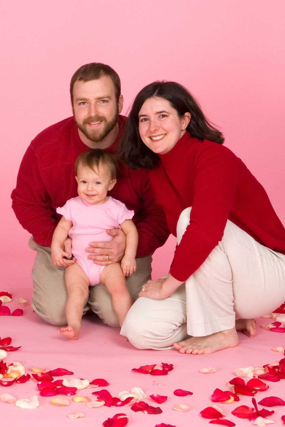 a family of mom, dad, and baby on a pink background surrounded by rose petals.